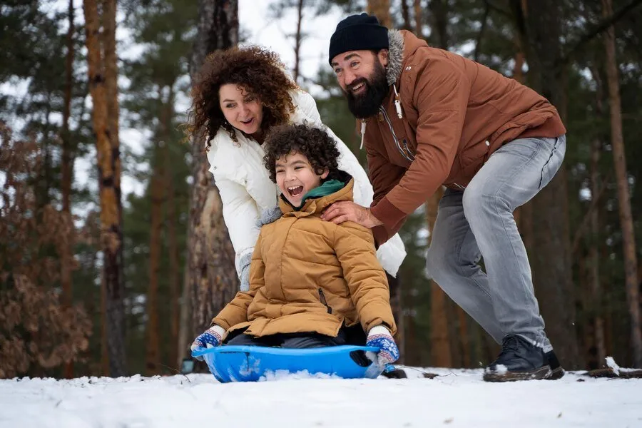 Moving-to-British-Columbia-with-kids Parents pushing a laughing child on a blue sled through a snowy forest