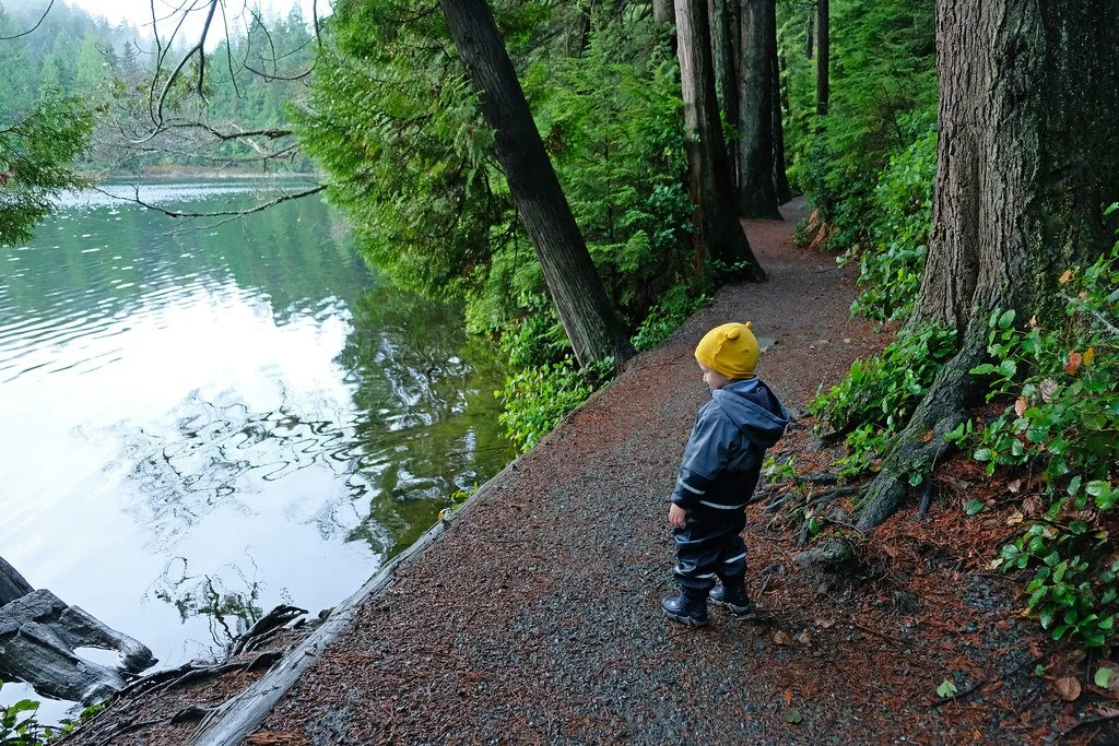 Moving-to-British-Columbia-with-kids A young child in a yellow hat standing on a forest trail beside a lake