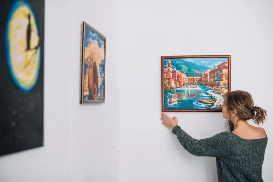 Prominent-Art-Galleries A woman hanging a framed landscape painting on a gallery wall