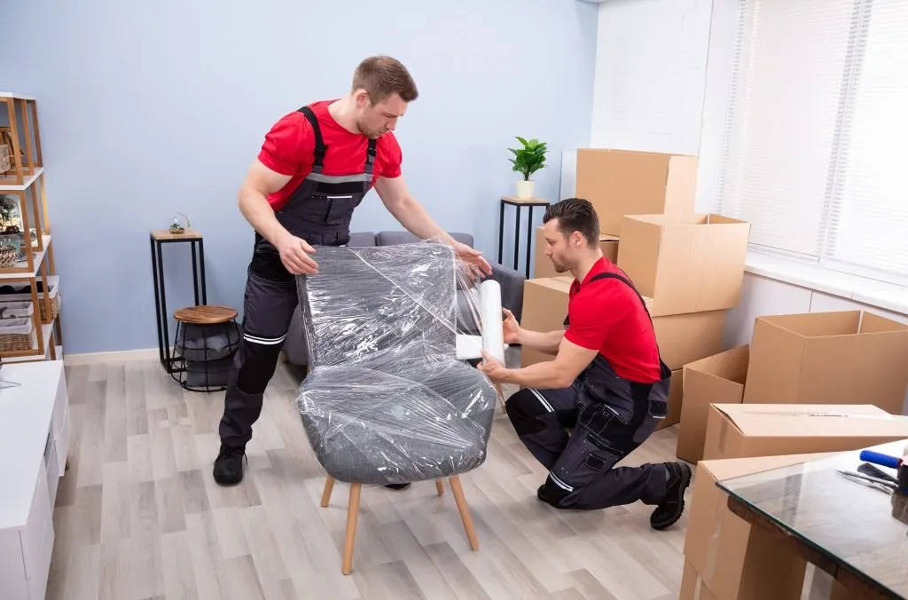 Two movers wrapping an armchair in protective plastic film beside stacked cartons