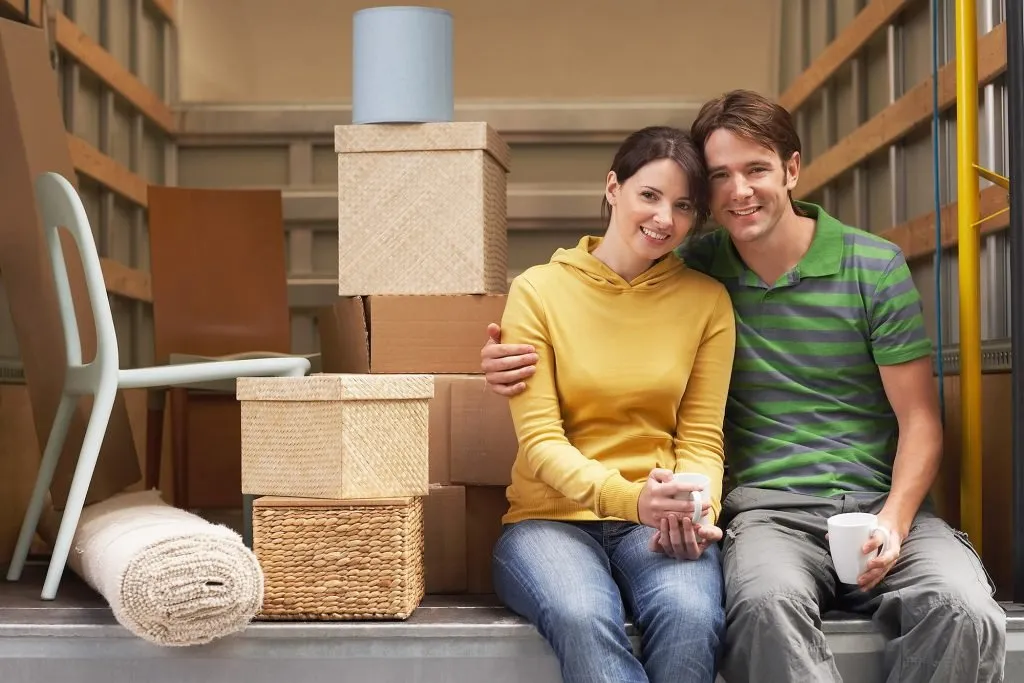 A couple sitting in a moving truck with mugs of coffee beside their packed belongings
