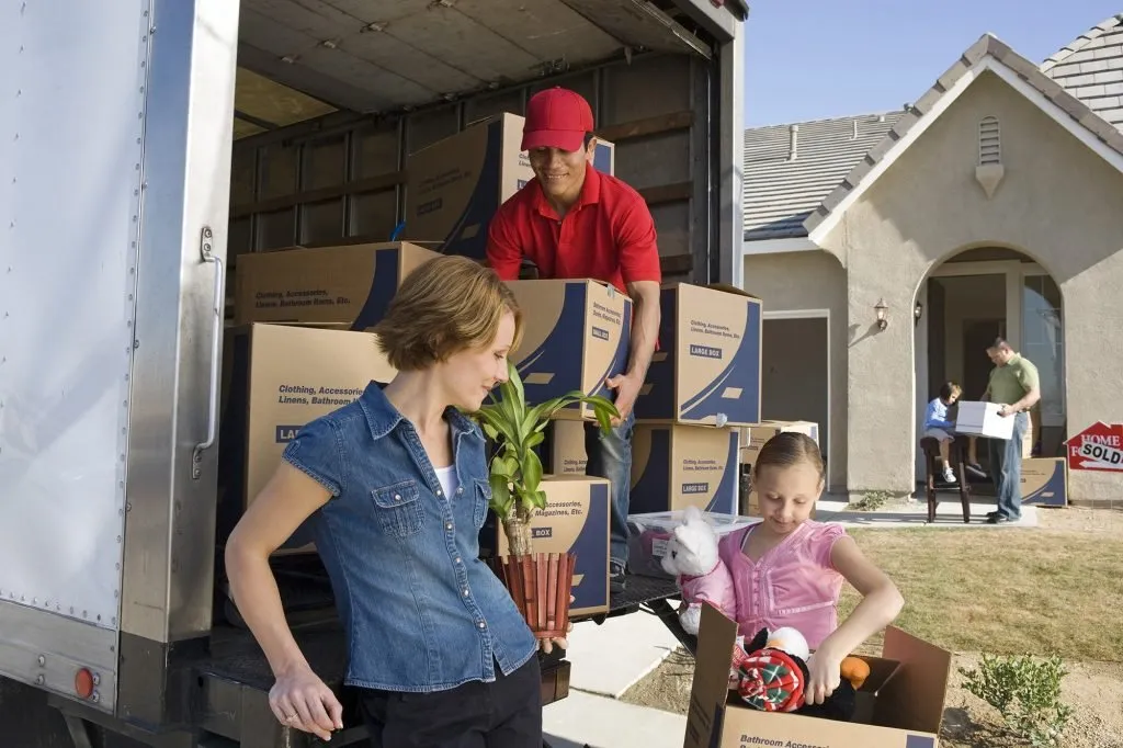 A family unloading labelled cartons and a plant from a moving truck outside a house with a Sold sign