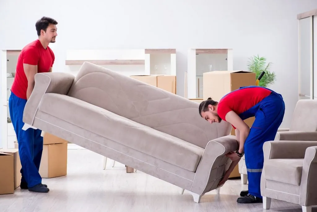 sofa-removals Two movers lifting a beige sofa in a living room surrounded by packed cartons