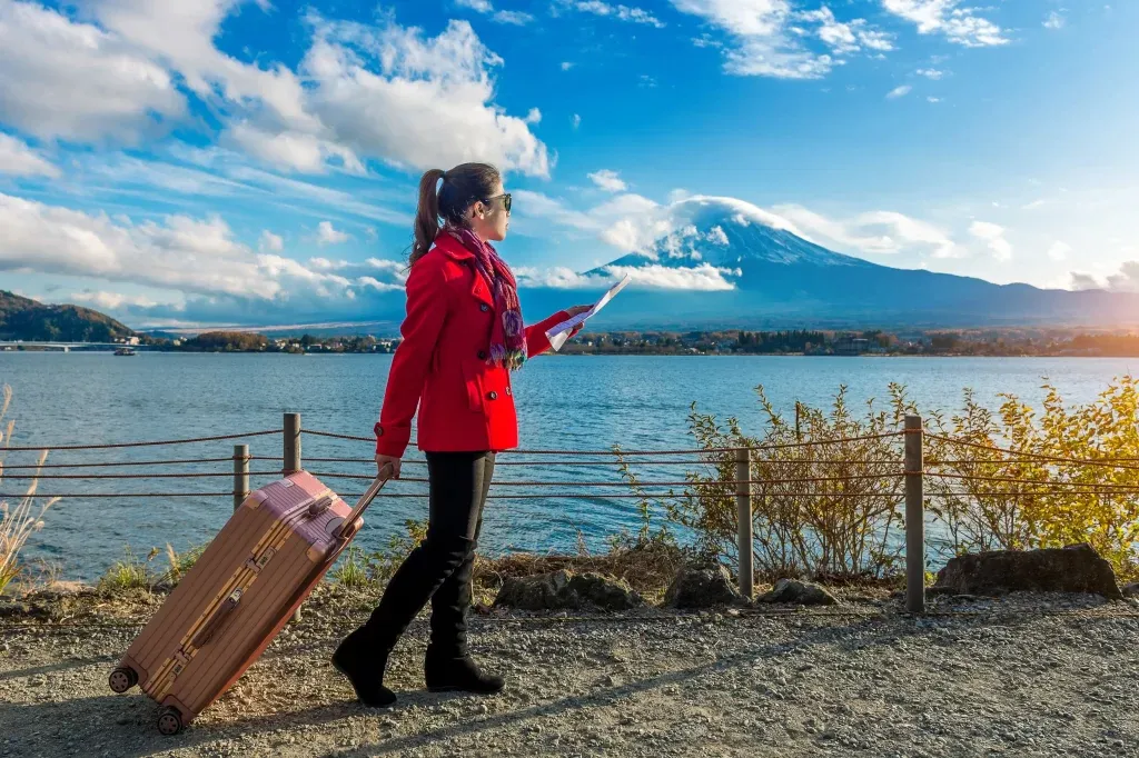 stay-safe-enjoy-British-Columbia A traveller in a red coat pulling a suitcase and reading a map beside a lake and snow-capped mountain