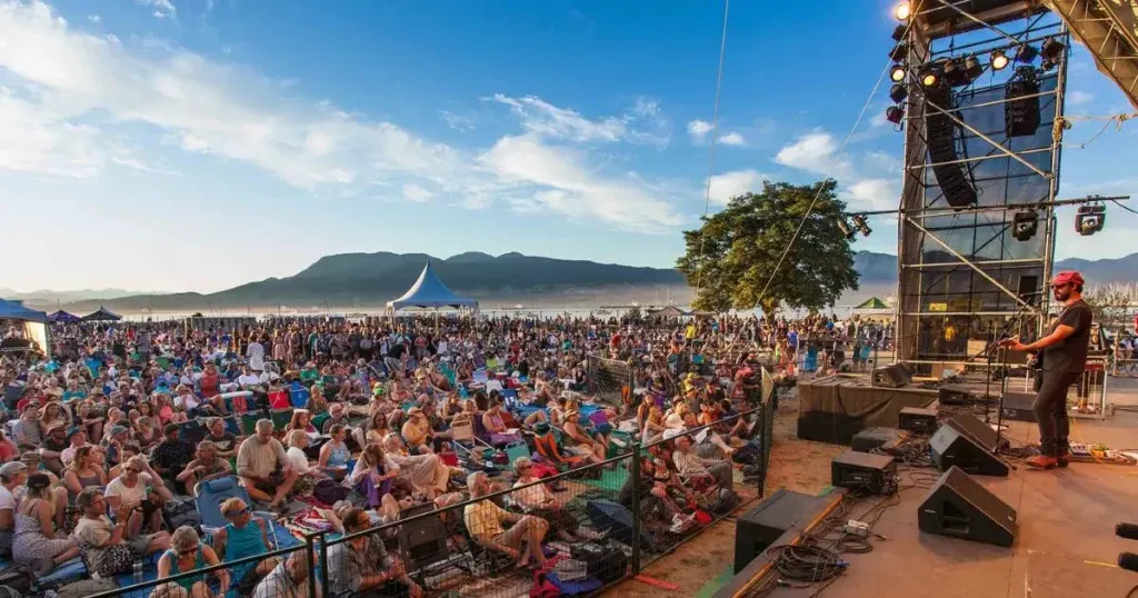 vancouver-folk-music-festival A seated crowd watching a performer on an outdoor festival stage with mountains behind